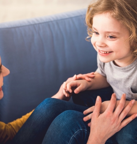 Mom and kid are grinning and looking into each-other eyes while staying down on sofa. They are joyfully hand-holding and boy is sitting on parent feet. Mother and child together concept