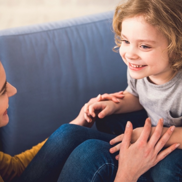 Mom and kid are grinning and looking into each-other eyes while staying down on sofa. They are joyfully hand-holding and boy is sitting on parent feet. Mother and child together concept