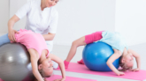Two smiling girls stretching their back on exercise balls, instructed by a physiotherapist