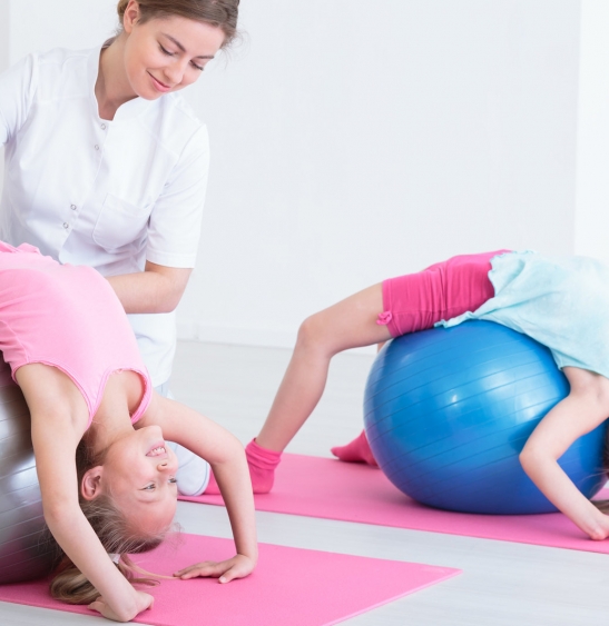 Two smiling girls stretching their back on exercise balls, instructed by a physiotherapist